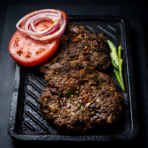 Two dark brown kabab patties with sliced tomatoes, red onions, and green chilies on a black tray.