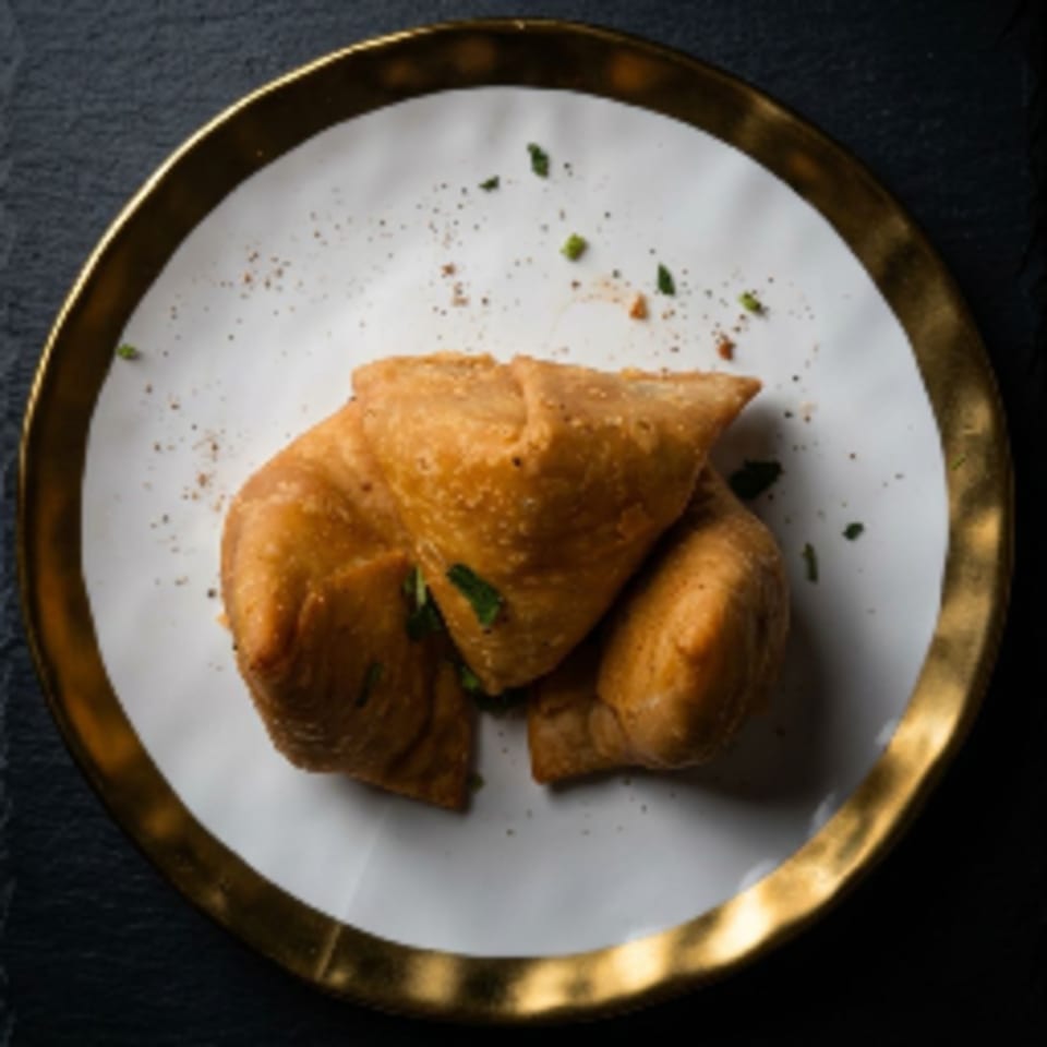 Three golden-brown samosas, garnished with herbs and spices, on a white plate with a gold rim.